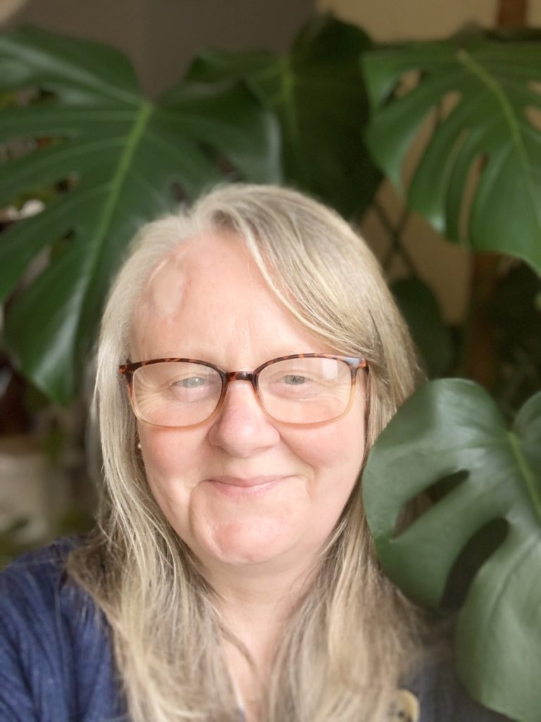 Sara Venn posing indoors next to a large monstera plant