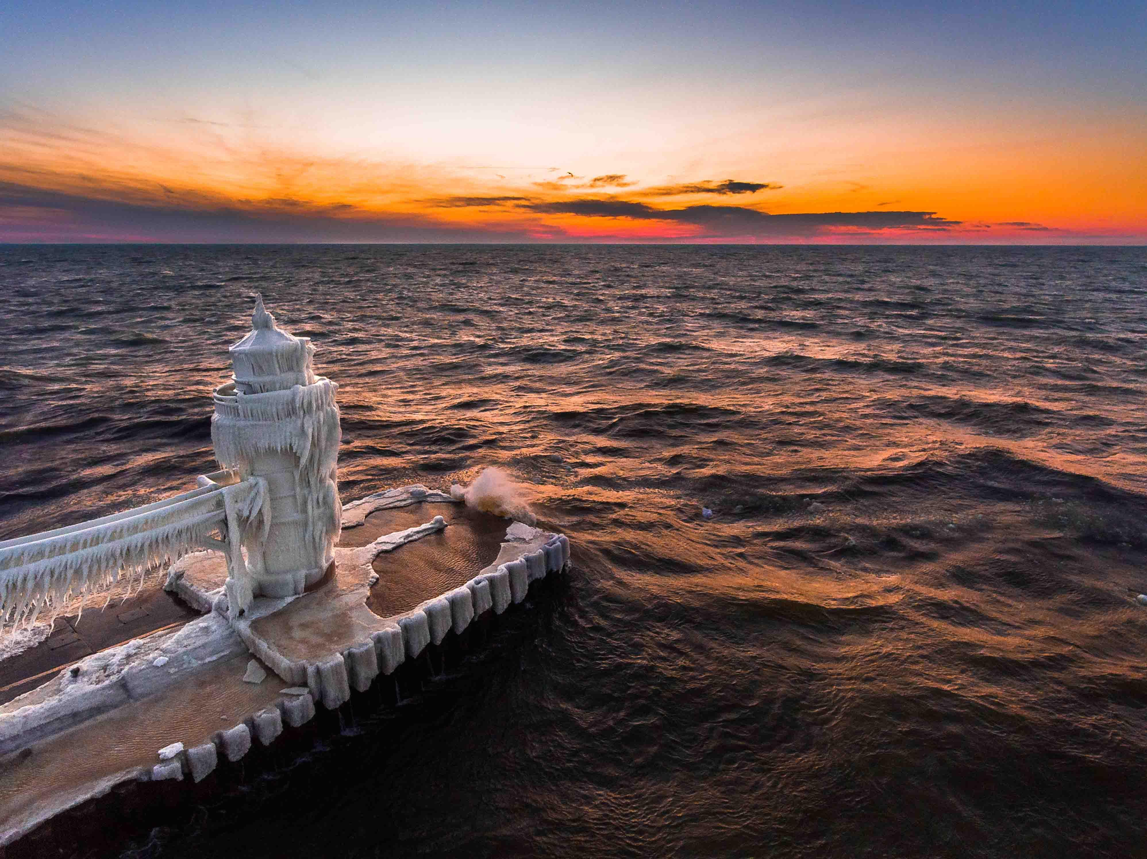 Waves crashing over a frozen lighthouse at sunset in St. Joseph