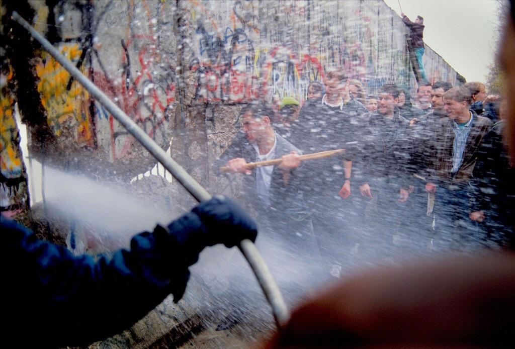 West German men hammer at the Berlin Wall while East German police blast water at them through