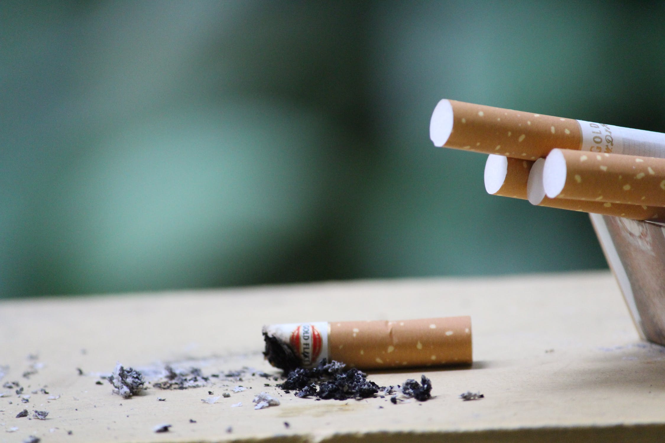 A photo of some cigarettes in an ashtray to the right. A cigarette butt is in the middle of the photo with ash on the table.