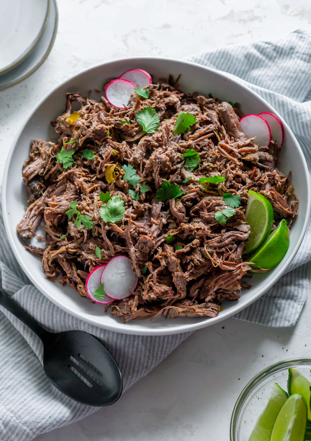 Beef barbacoa in a white bowl with limes and radishes.
