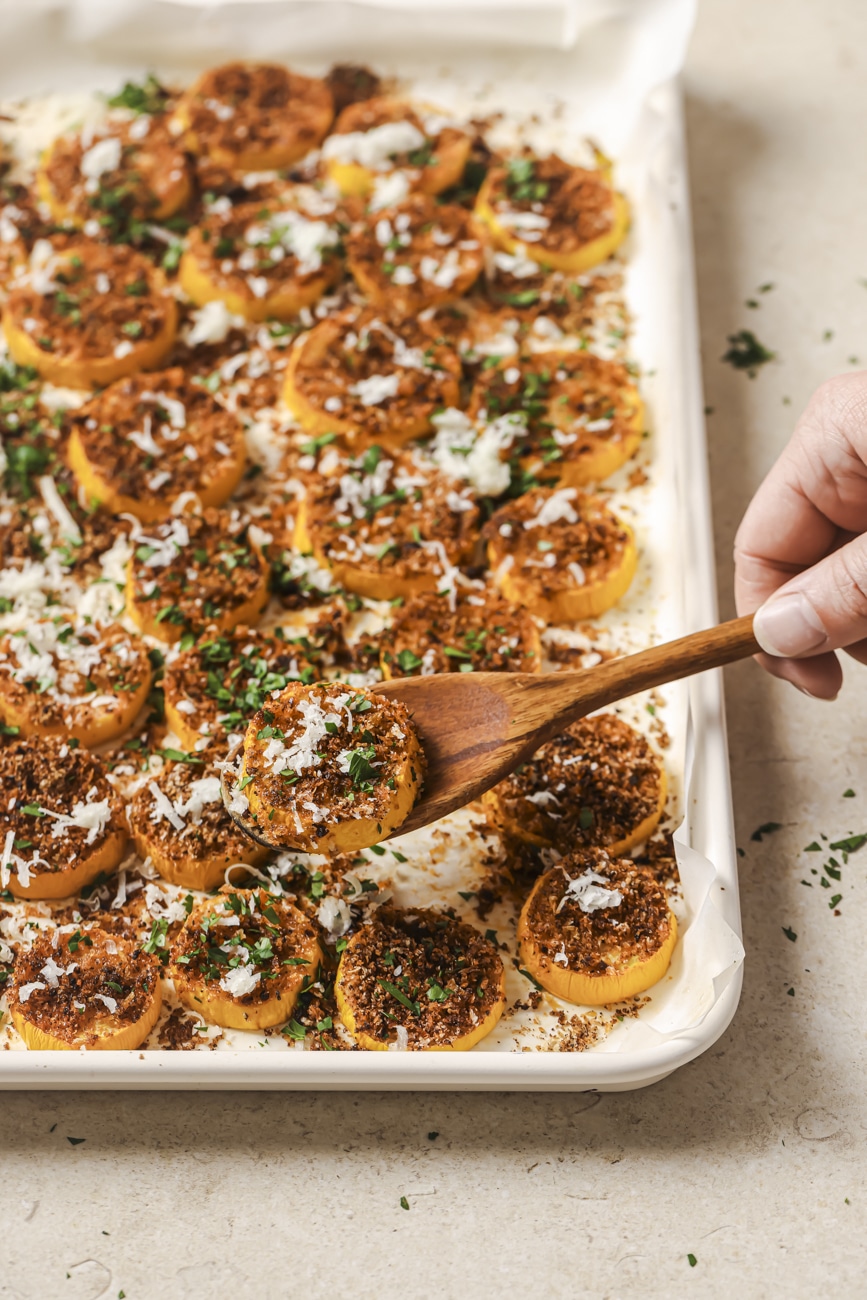 a wooden spoon holding a piece of roasted yellow squash over a sheet pan full of cooked squash.