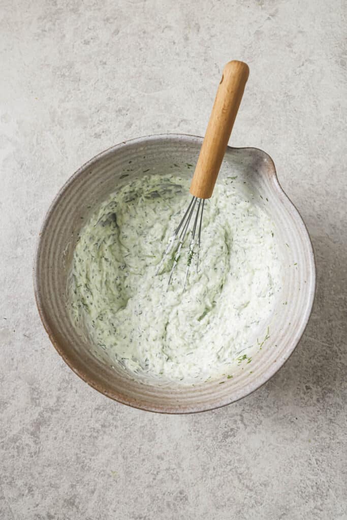ingredients for tzatziki sauce combined in a bowl with a whisk resting on the side