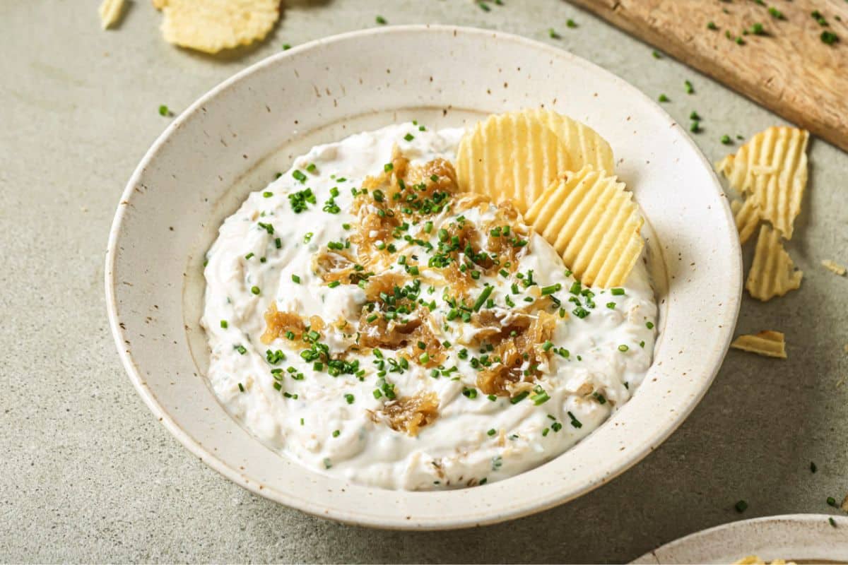 A white bowl filled with homemade french onion dip with a ruffled chip resting on top.