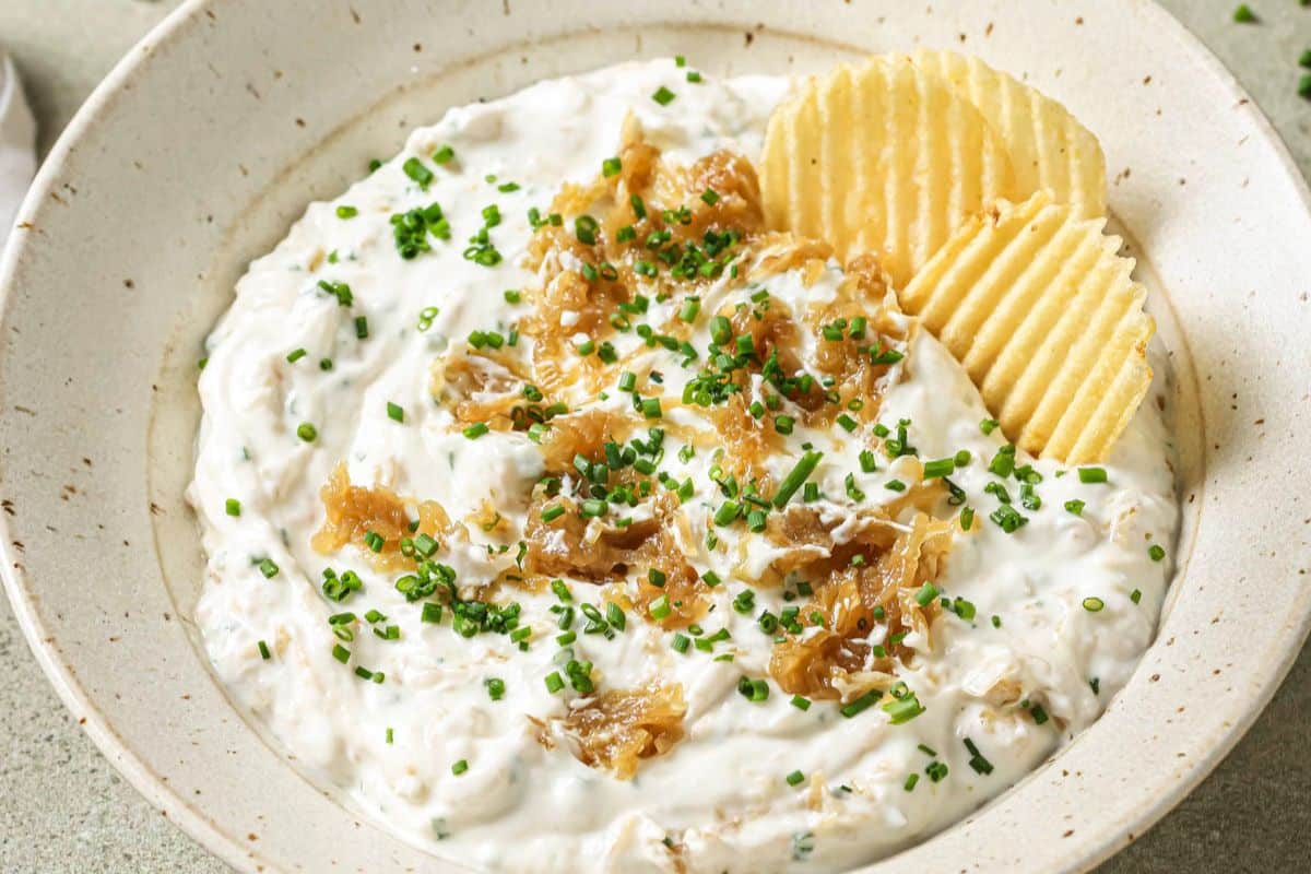 Up close shot of a homemade French onion dip recipe in a bowl topped with caramelized onions, fresh chives, and potato chips.