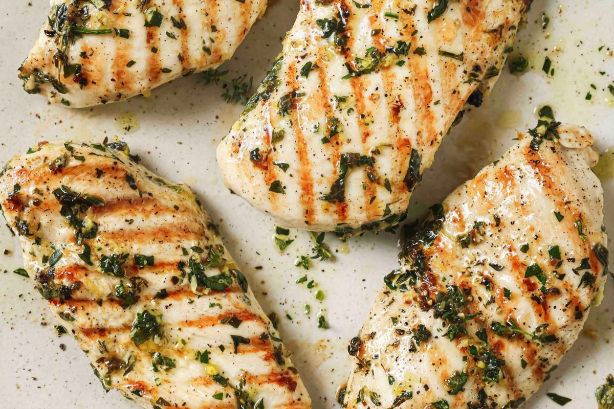 Four lemon herb grilled chicken breasts on a white counter topped with fresh herbs.