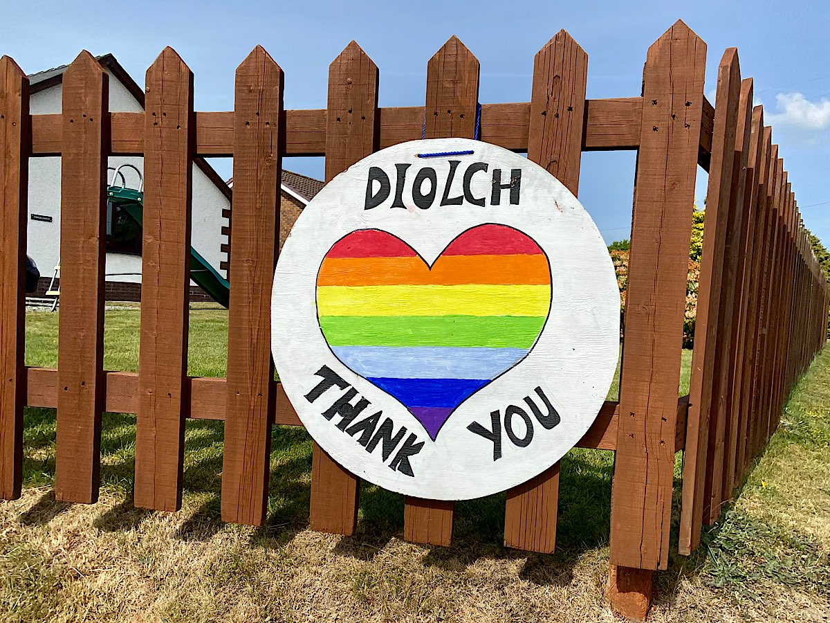 a big white circular board with a heart shaped painted in rainbow colours. Above the heart ir reads 'Diolch' and below the heart is reads 'Than You' - it's hanging on a very tidy brown picket style fence
