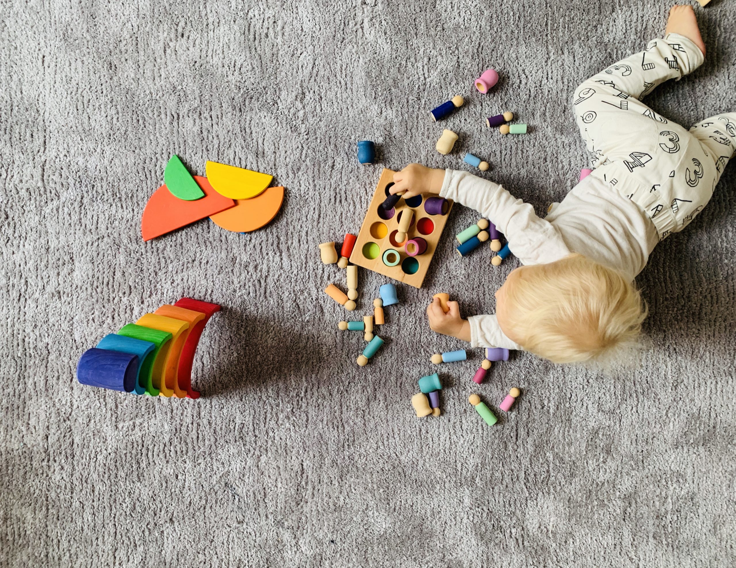 Lintuperspektiivist&auml; otettu kuva, jossa valkoihoinen ja vaaleahiuksinen lapsi leikkii v&auml;rikk&auml;ill&auml; palikoilla harmaan maton p&auml;&auml;ll&auml;. 