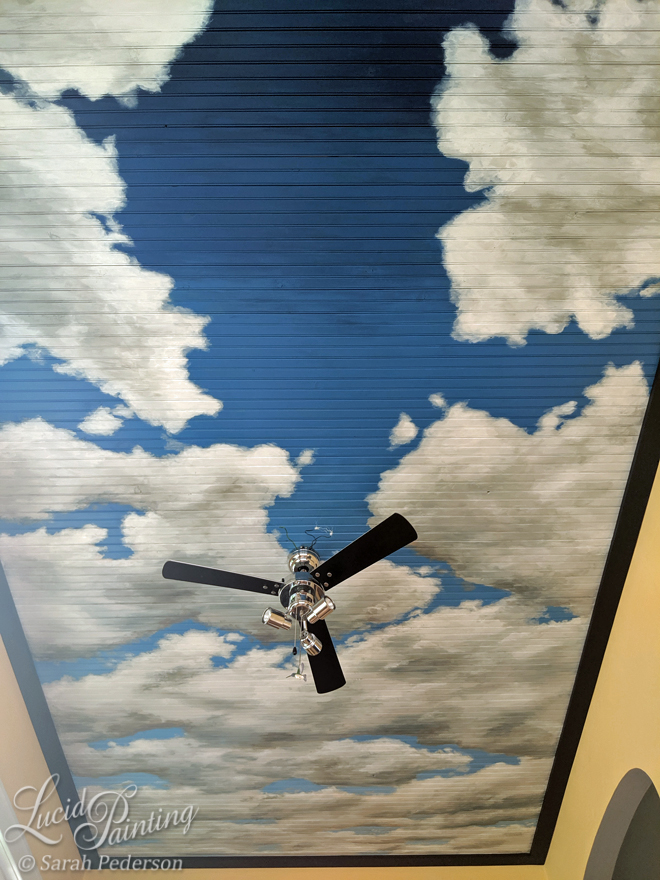 A blue sky mural with clouds, painted in perspective on a beadboard ceiling of a home office.