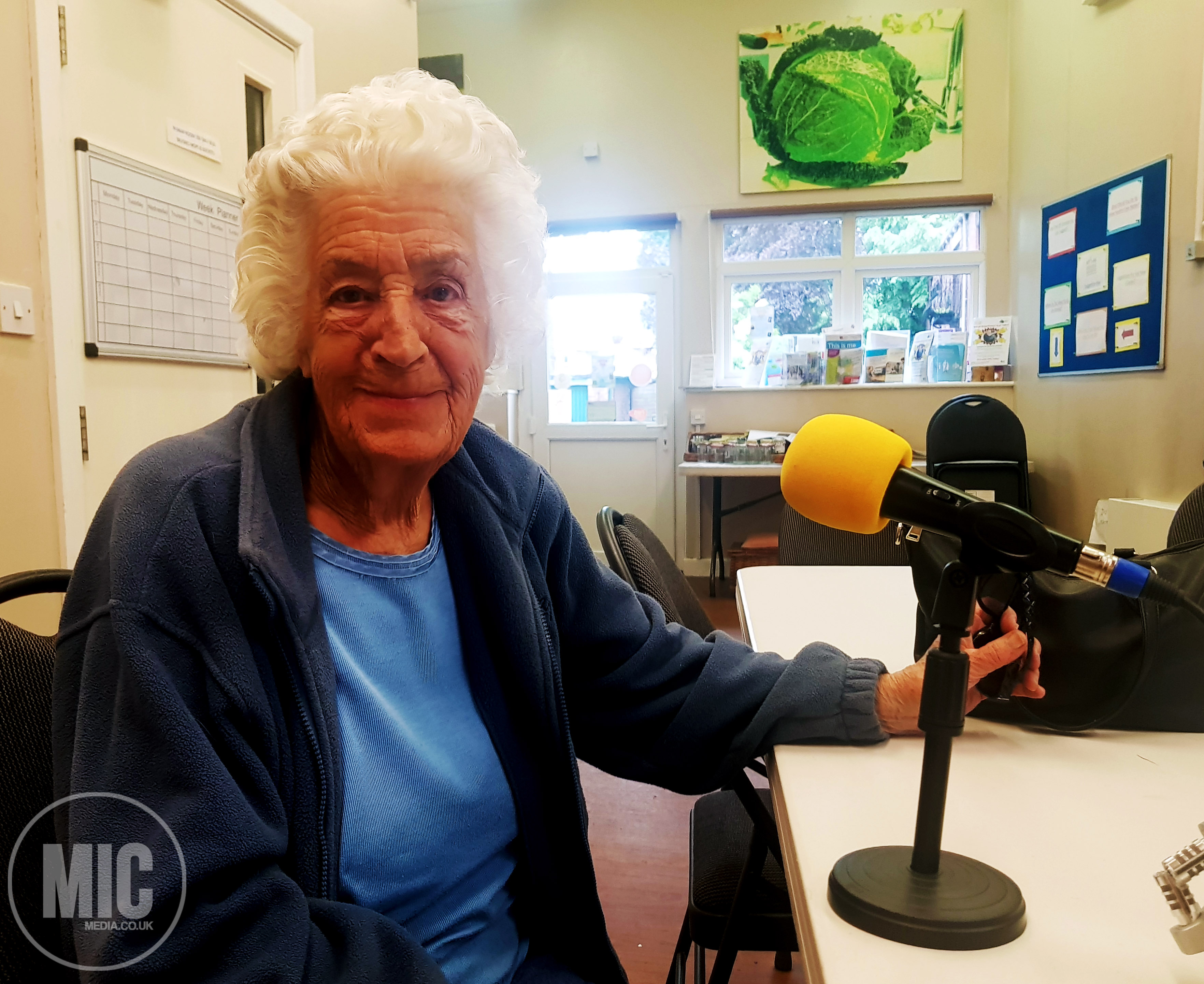 a photo of a women with white hair and a blue tshirt. she is sat on a chair. we can see her from the waist up. the3 table in front of her has a microphone with a yellow foam end on it. The microphone is pointed towards the lady