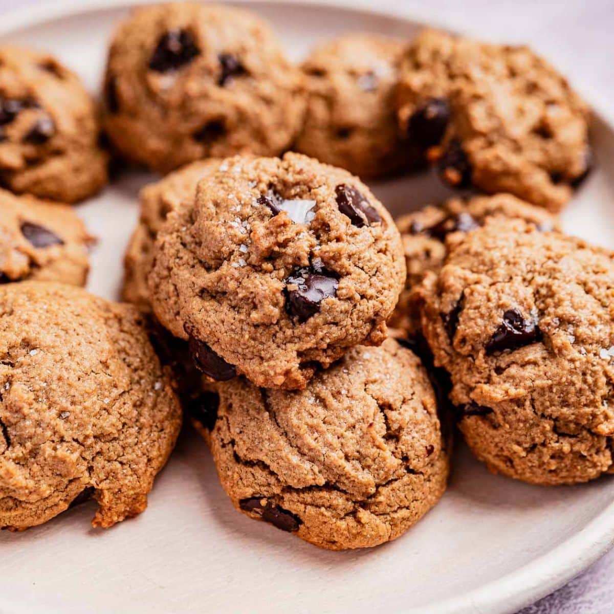 A plate of oat flour chocolate chip cookies rests on a white surface, showcasing a light brown texture with visible chocolate chunks.