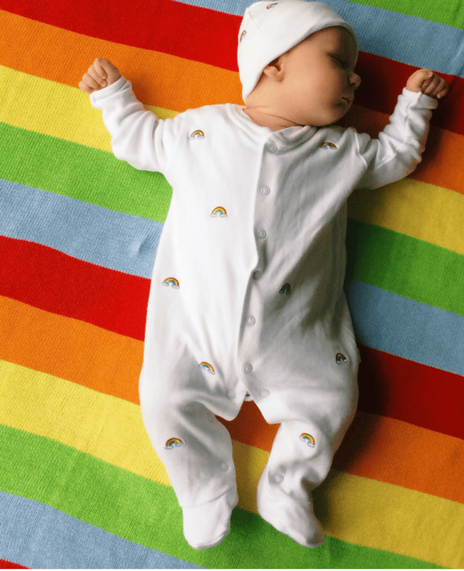A gorgeous baby boy asleep on a brightly coloured rainbow blanket - he is wearing a Jojo Maman Bebe sleepsuit and matching hat - they are white and covered in tiny embroidered rainbows - Little Mister H at two months old - Mrs H's favourite things