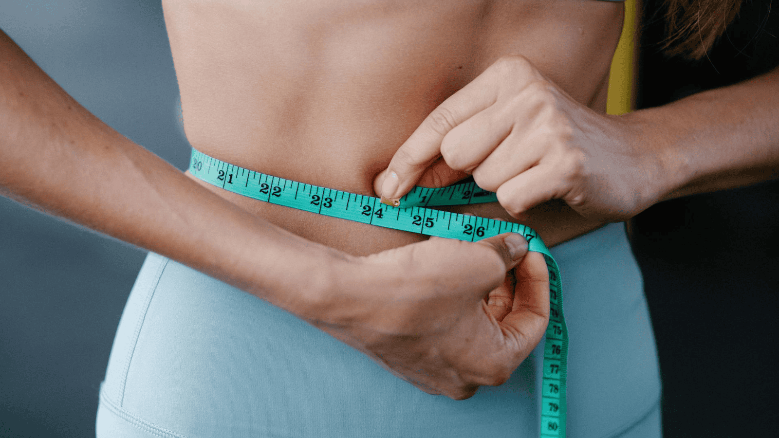 A person's hands holding a blue measuring tape around their bare waist, while wearing light blue athletic pants. Only the torso is visible, and the background is out of focus.