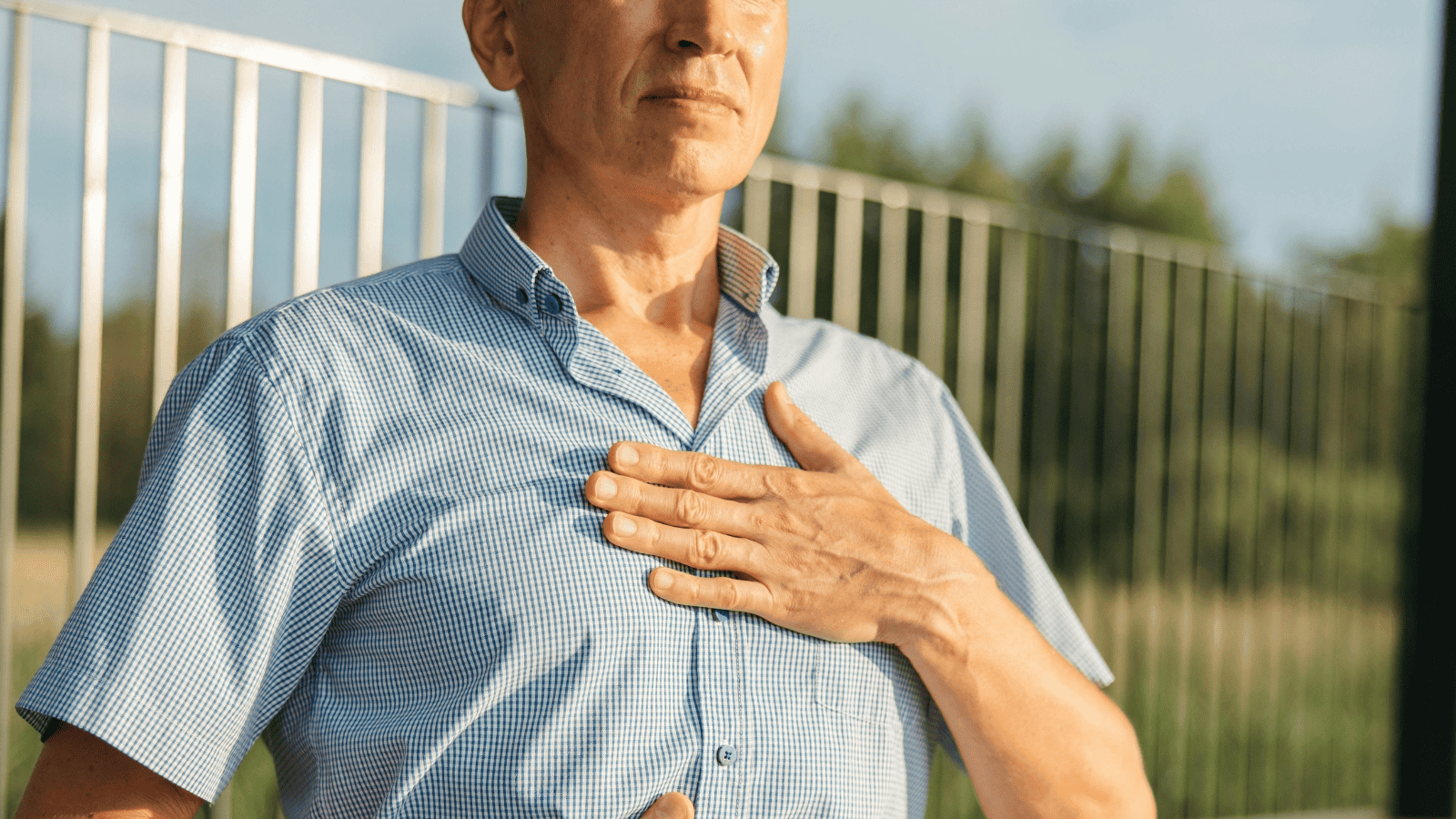 A person in a blue checkered shirt holds their hand to their chest while standing outside near a metal fence. The background features blurred trees and a blue sky.