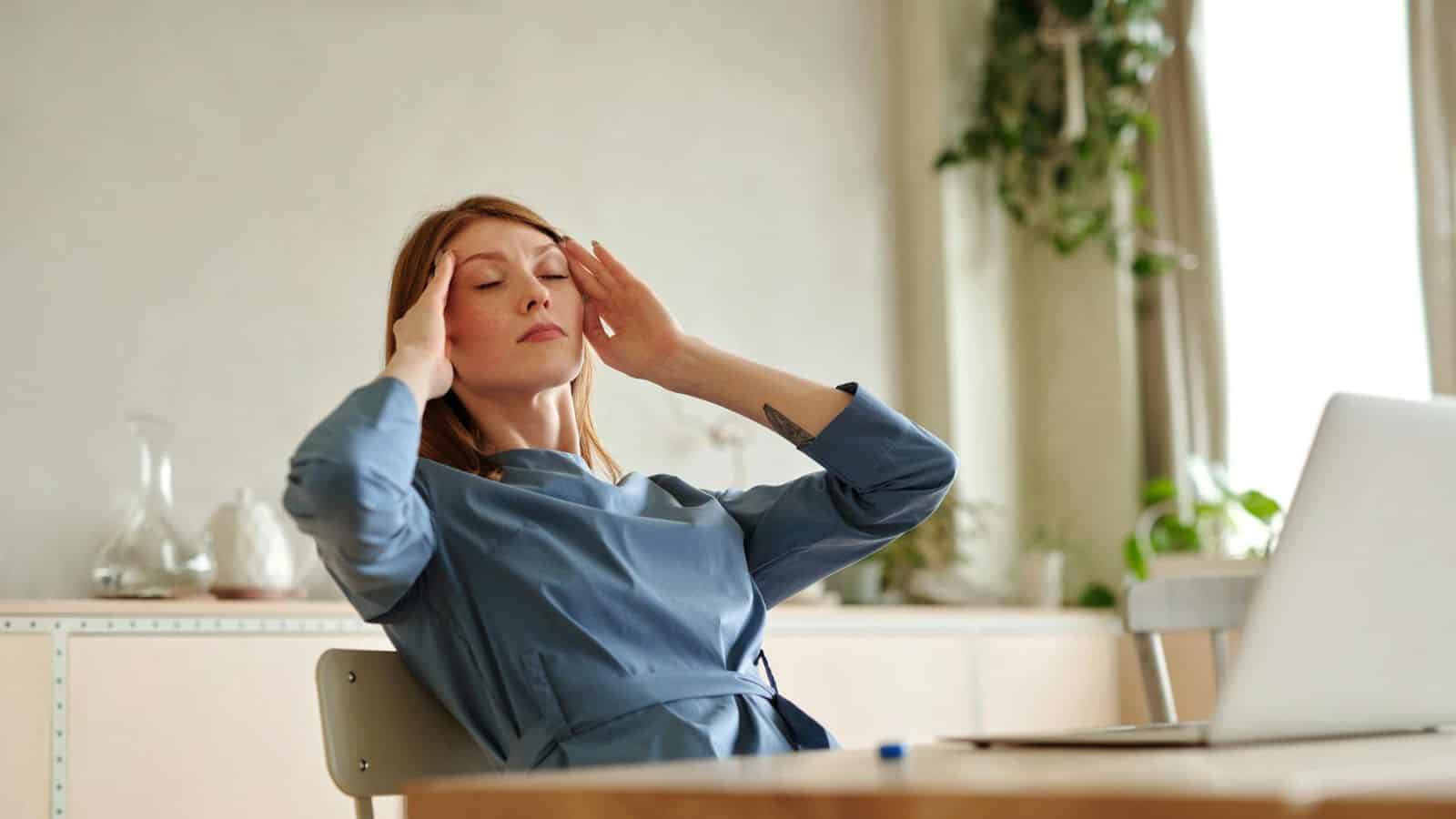 Woman with both hands on temples while sitting down in front of her laptop