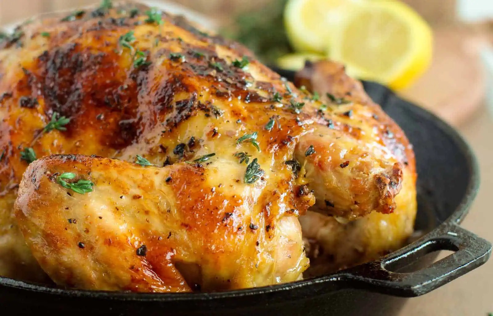A whole roasted chicken seasoned with herbs sits in a skillet, garnished with fresh thyme. Lemon slices are visible in the background.