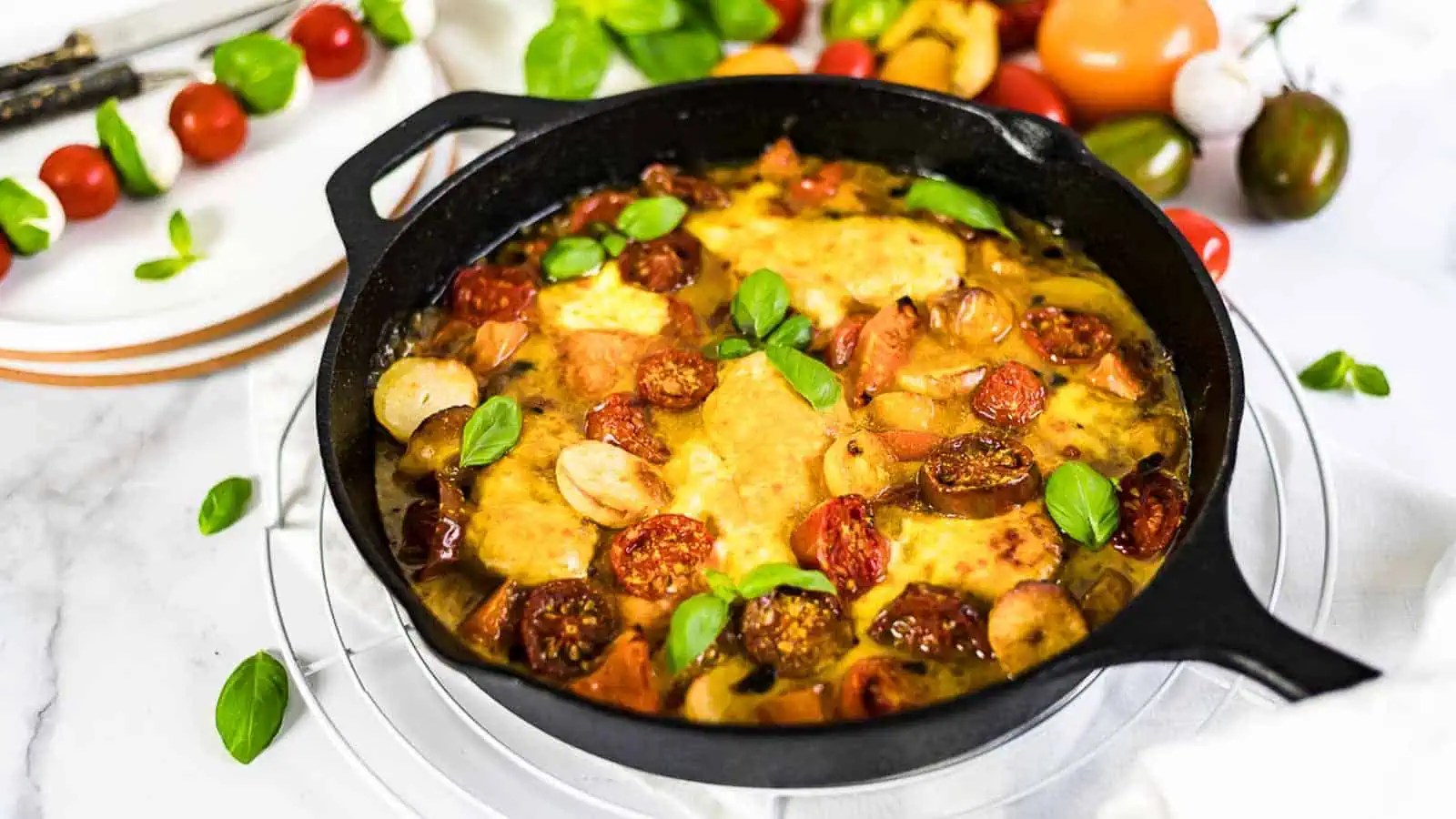 A cast iron skillet filled with a baked dish featuring chicken, sliced tomatoes, sausage, and topped with fresh basil leaves, surrounded by tomatoes and basil on a marble surface.