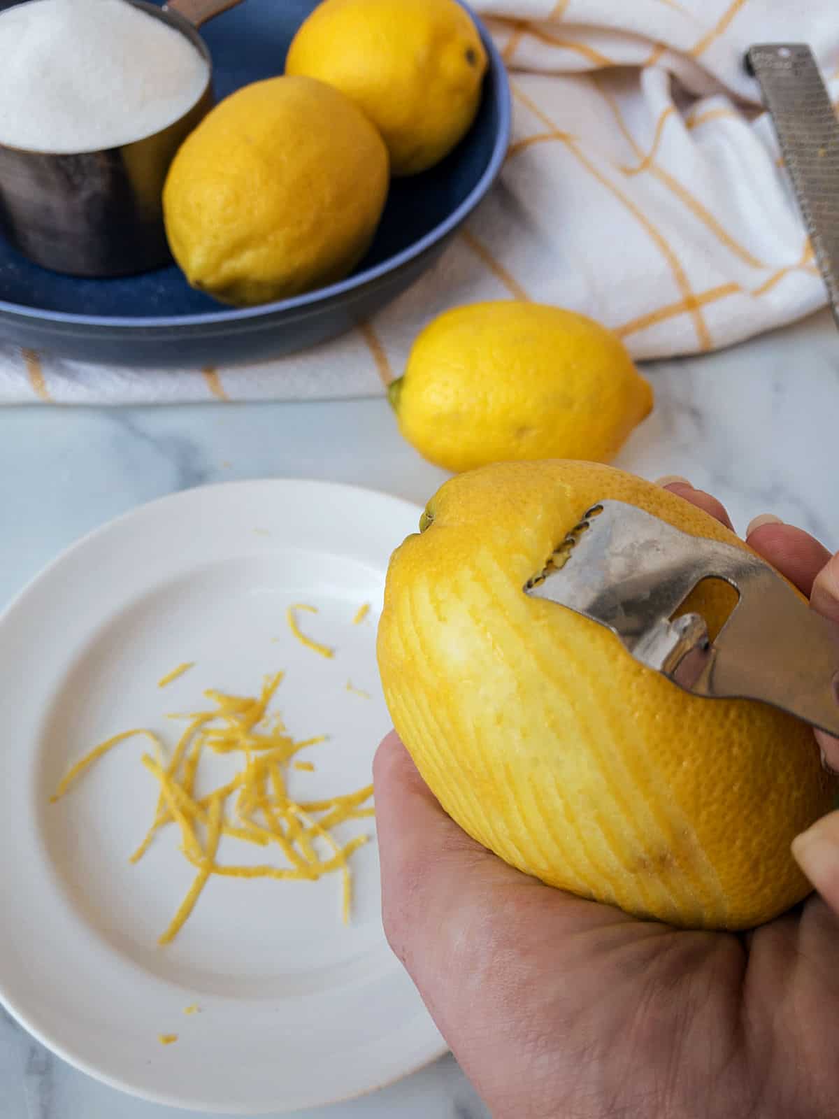 A person is zesting a lemon with a metal zester over a white plate.