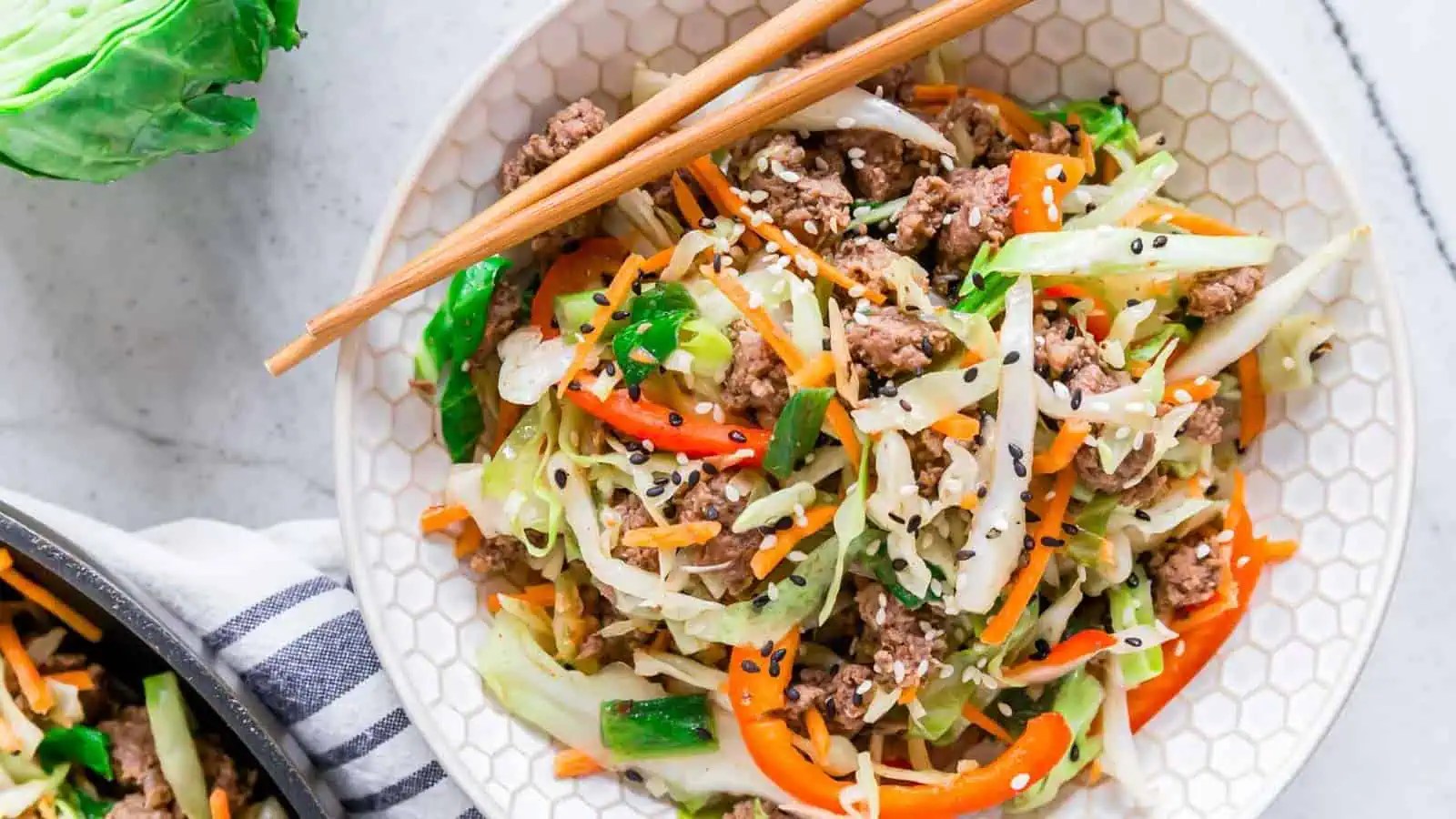 A bowl of stir-fried vegetables and ground meat garnished with sesame seeds, with a pair of chopsticks resting on top.