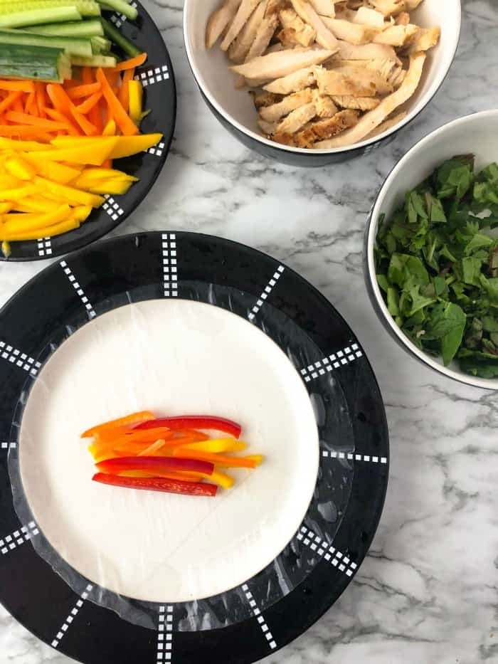 A spring roll rice wrapper on a plate with peppers in the middle, other chopped ingredients in bowls next to the plate