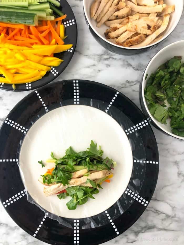 A fresh spring roll wrapper on a plate with chicken and greens in the roll, other ingredients in bowls next to the plate.