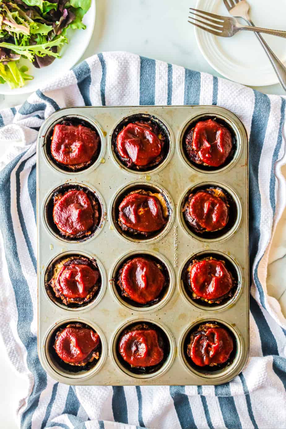 Mini meatloaf in a muffin pan from overhead topped with bbq sauce with salad and plates in the background