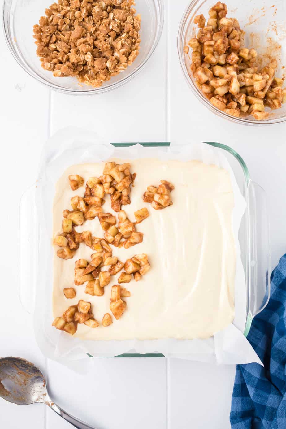 Layering the apple layer on top of the cheesecake layer in a square pan from overhead.