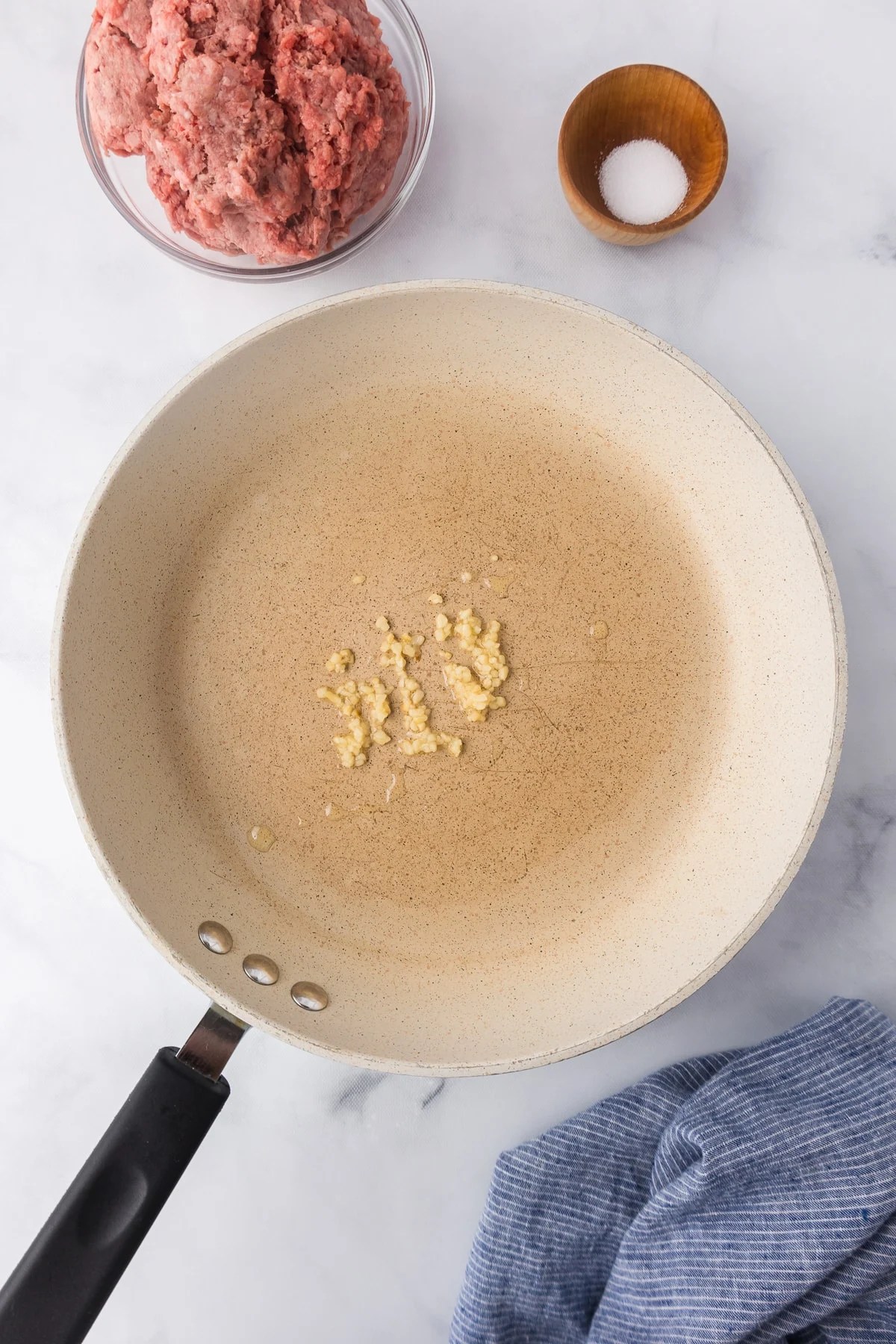A pan with garlic in the pan, with raw ground beef and salt in bowls nearby.