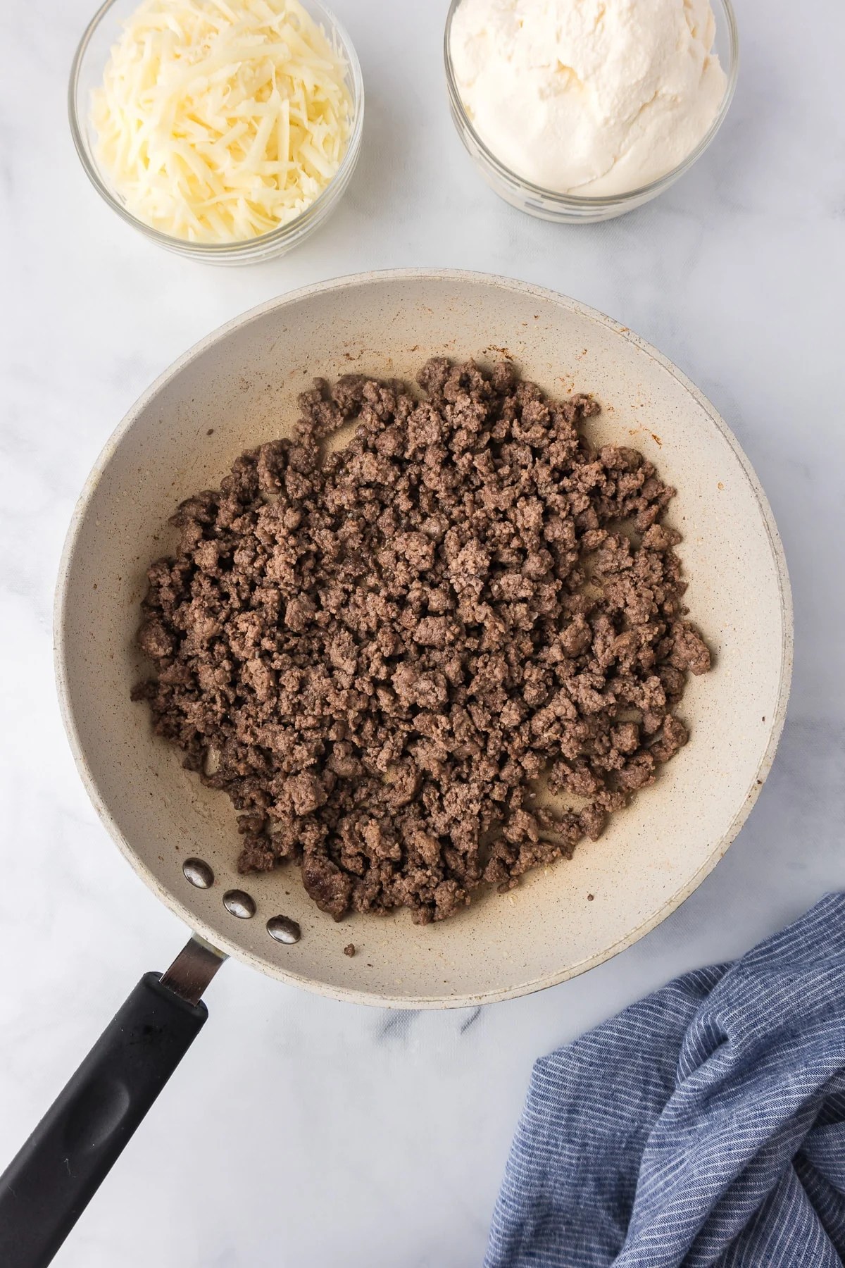 A frying pan with cooked ground beef with parmesan cheese and ricotta cheese in bowls nearby on a kitchen counter.