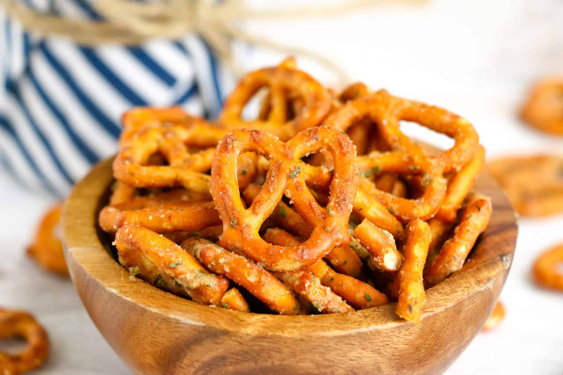 A wooden bowl filled with garlic ranch pretzels sits on a white surface, with a blurred striped cloth in the background.