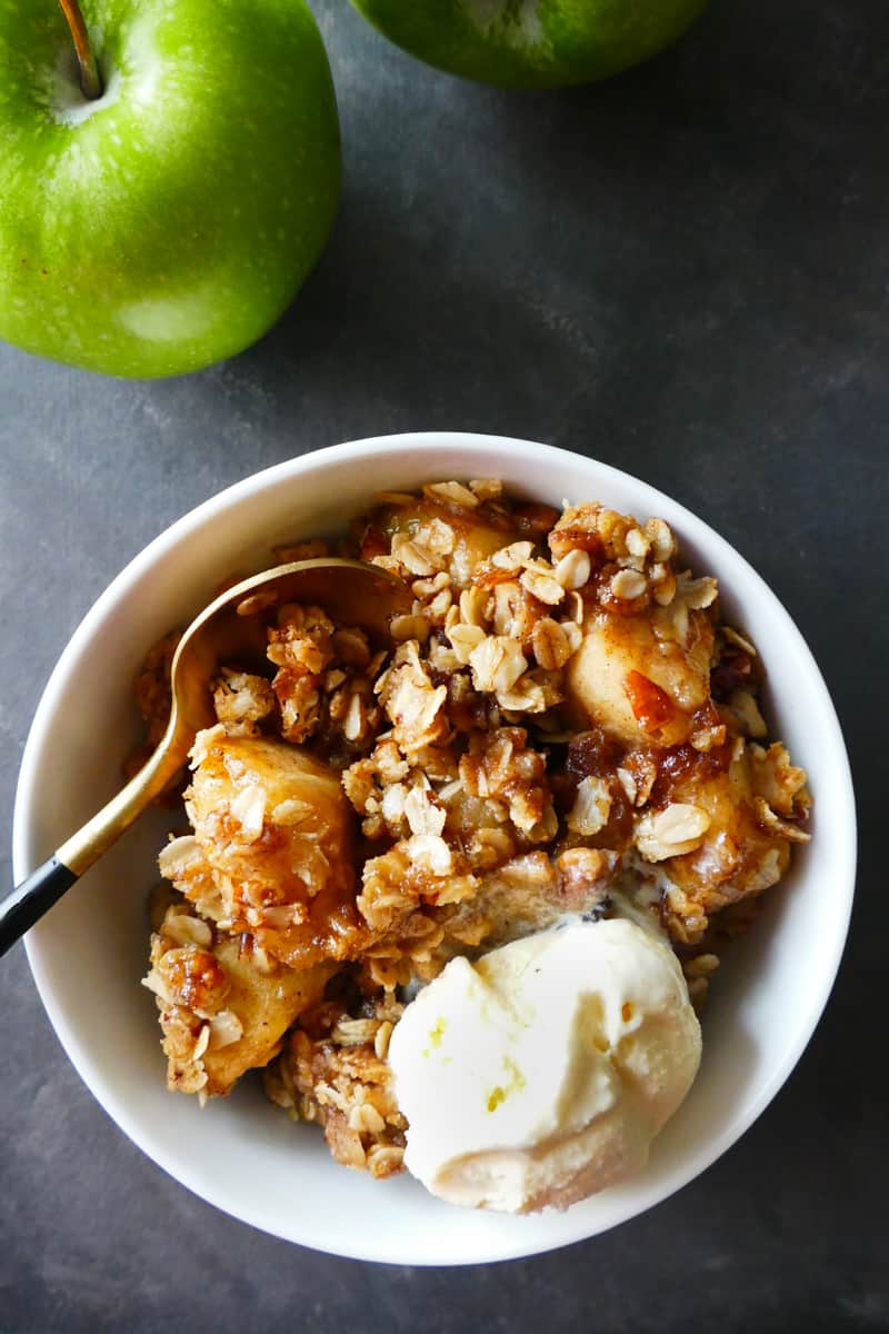 White bowl with apple crisp and scoop of ice cream with a gold spoon nestled in the bowl and green apple in the background