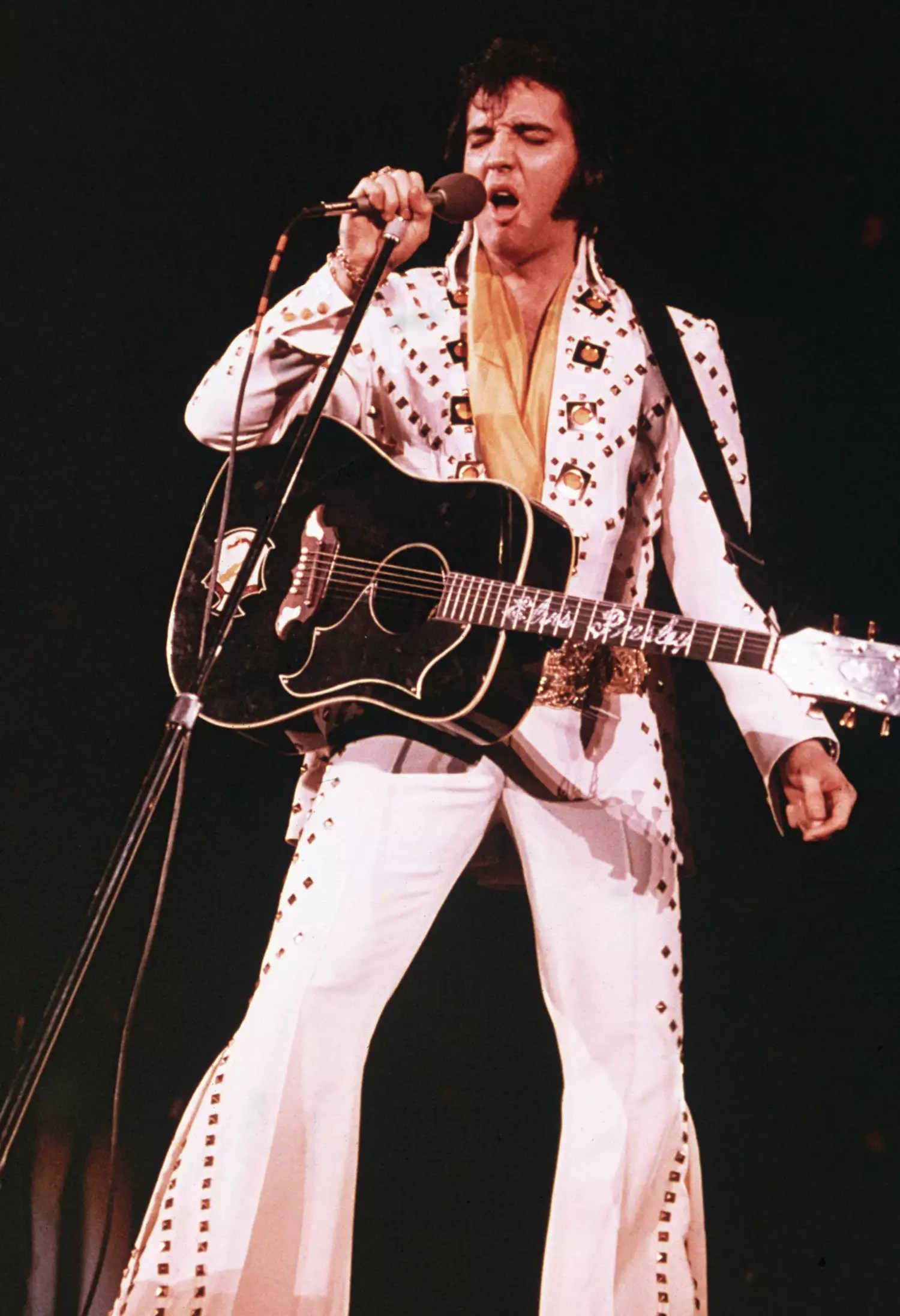 Elvis Presley (1935 - 1977), wearing a white rhinestone-studded suit and strapped guitar, singing into a microphone with his eyes closed.