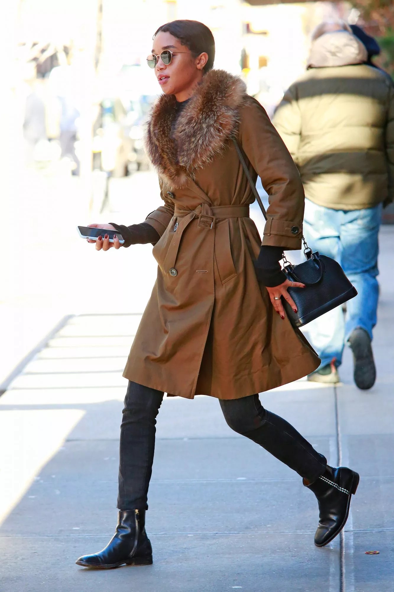 EXCLUSIVE: Laura Harrier Wears A Brown Trench Coat With Fur Accents Paired With Black Pants, Boots And Louis Vuitton Shoulder Bag