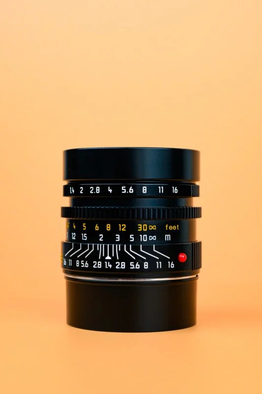 A black camera lens with white and yellow aperture and focus markings stands upright on an orange surface, against a matching orange background.