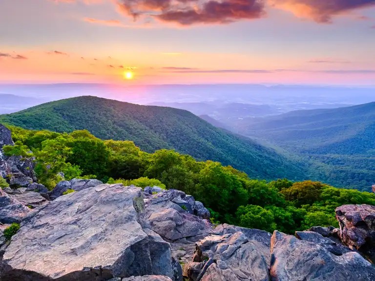 View from a mountain summit looking out over rolling forests under a purple and orange sky at sunset.