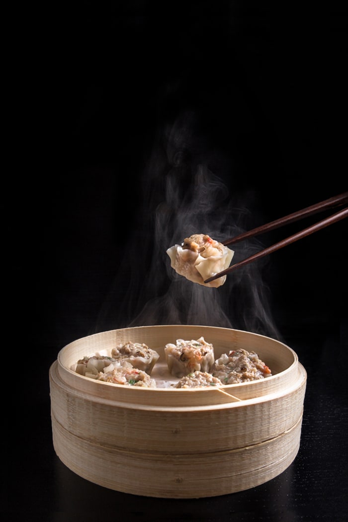 Shumai (Shrimp & Pork Dumplings) in a steamer basket. One dumpling being held with chopsticks