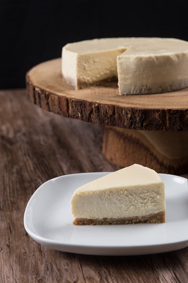 a slice of New York Cheesecake made in a pressure cooker on a white plate with rest of cheesecake in the background on a tree trunk syle cake stand