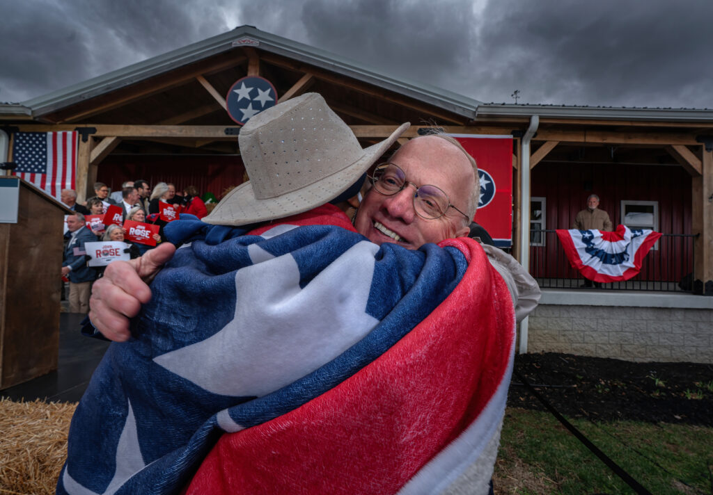 Through A Glass Darkly: The Photographs That Told 2025’S Stories 12 A woman wearing a cowboy hat and wrapped in a Tennessee flag hugs a balding man with glasses.