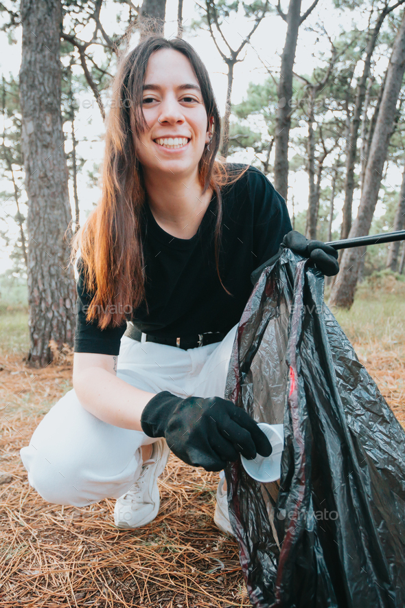 A young woman collects abandoned garbage in a black bag in the forest