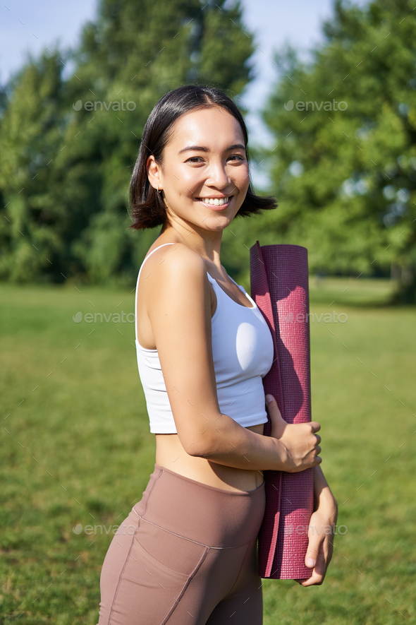 Smiling fitness girl with rubber mat, stands in park wearing uniform