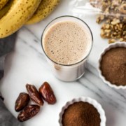 A glass of smoothie with banana. walnuts, coffee powder and dates next to it