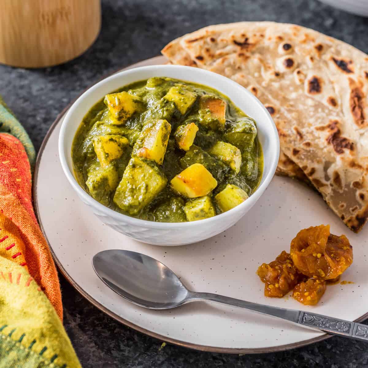 A white plate with a white bowl filled with aloo saag on the left of the plate and a silver spoon at the front of the plate with fresh paratha in the back of the plate.