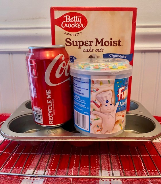 A box of chocolate cake mix, a can of cola, and a tub of frosting are shown sitting atop of a cupcake tray.