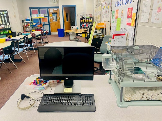 A black computer monitor is shown on a table in a classroom. There is a keyboard and a pair of headphones next to the computer.