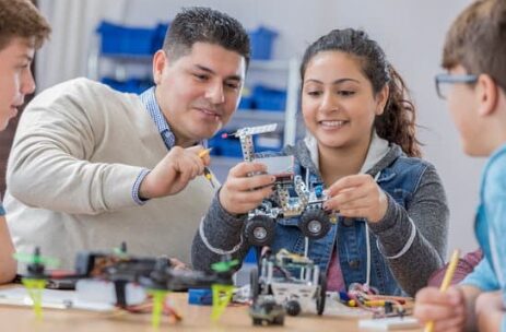 A teacher and students are seated at a table, working on a robotics project.