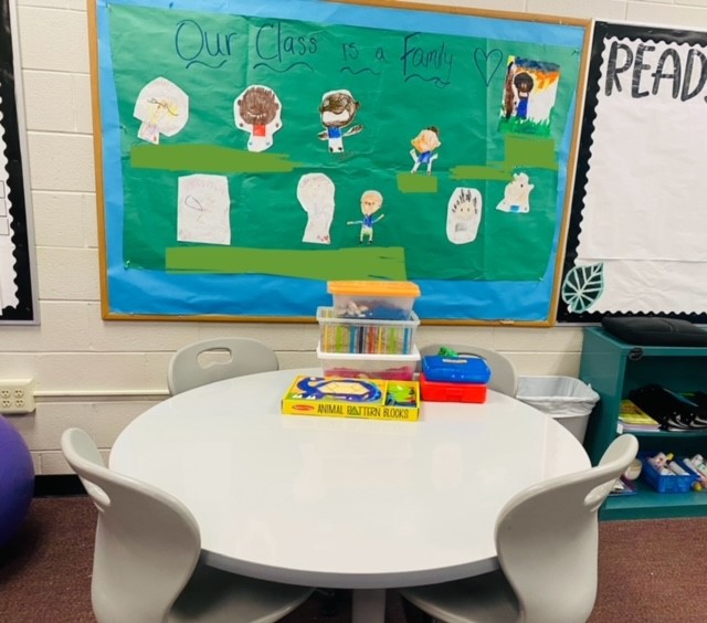 A round white table with chairs in front of a bulletin board. There are some bins and toys on the table.