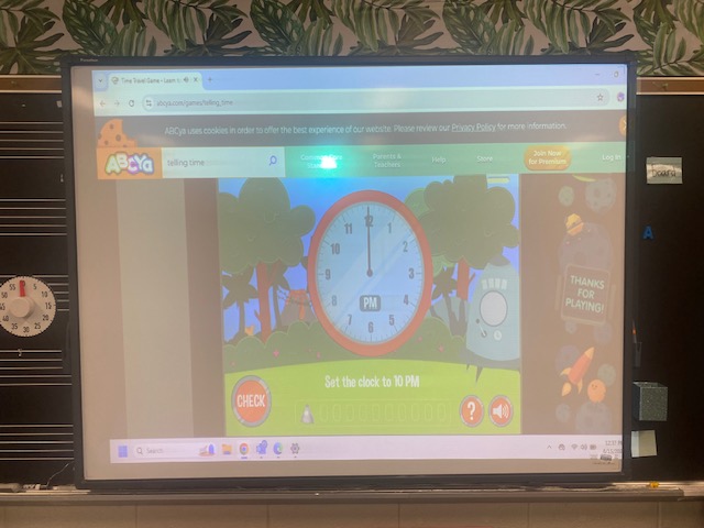 A Smartboard is shown in a classroom. On the screen is a game showing an analog clock.
