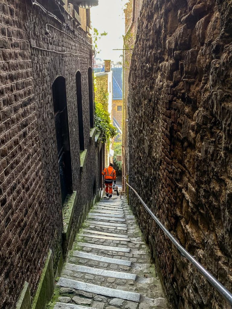 Lüttich, Impasse de Ursulines nach unten fotografiert, am Fuße ist ein Straßenkehrer, der mit einem Laubbläser Blätter von den Stufen fegt.