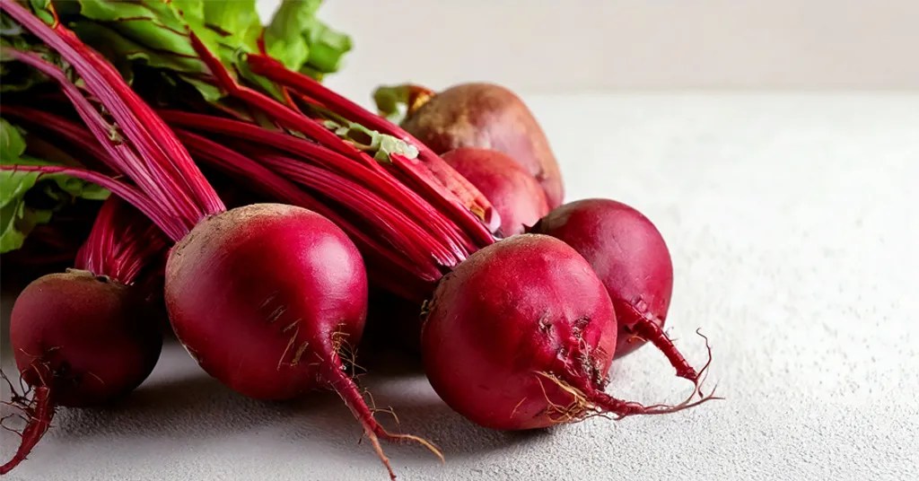 Freshly harvested red beets with leafy tops and long magenta stems arranged on a white surface, showcasing their vibrant color and farm-to-table appeal.