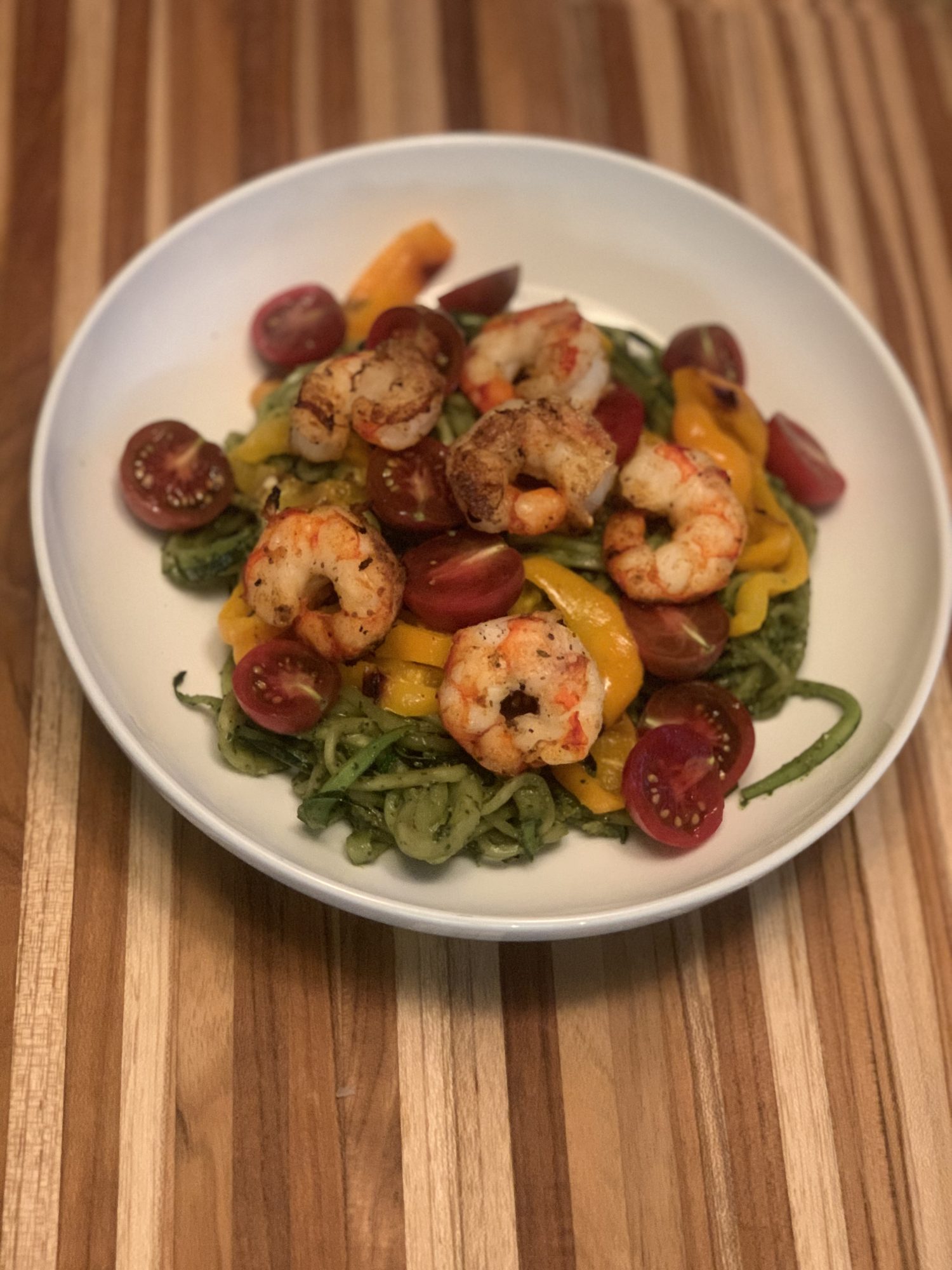 A beautiful wood cutting board holding a white bowl full of pesto zucchini noodles, sliced cherry tomatoes, grilled shrimp and bell peppers. For more incredible recipes be sure to follow www.sprouting-vitality.com #sproutingvitality #kenzinthekitch @sproutingvitality